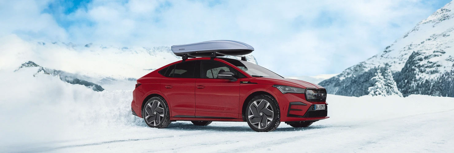 Das Skoda Enyaq Coupé mit einem Dachgepäckträger steht auf einer verschneiten Straße in einer bergigen Landschaft. Im Hintergrund sind schneebedeckte Gipfel und ein blauer Himmel mit Wolken zu sehen. Das Skoda Enyaq Coupé mit einem Dachgepäckträger steht auf einer verschneiten Straße in einer bergigen Landschaft. Im Hintergrund sind schneebedeckte Gipfel und ein blauer Himmel mit Wolken zu sehen.