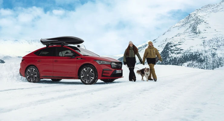 Das Skoda Enyaq Coupé in Rot mit einem Dachgepäckträger steht auf einer verschneiten Straße in einer bergigen Landschaft. Zwei Personen in Winterkleidung gehen mit einem Hund neben dem Fahrzeug. Im Hintergrund sind schneebedeckte Gipfel und ein blauer Himmel mit Wolken zu sehen. Das Skoda Enyaq Coupé in Rot mit einem Dachgepäckträger steht auf einer verschneiten Straße in einer bergigen Landschaft. Zwei Personen in Winterkleidung gehen mit einem Hund neben dem Fahrzeug. Im Hintergrund sind schneebedeckte Gipfel und ein blauer Himmel mit Wolken zu sehen.