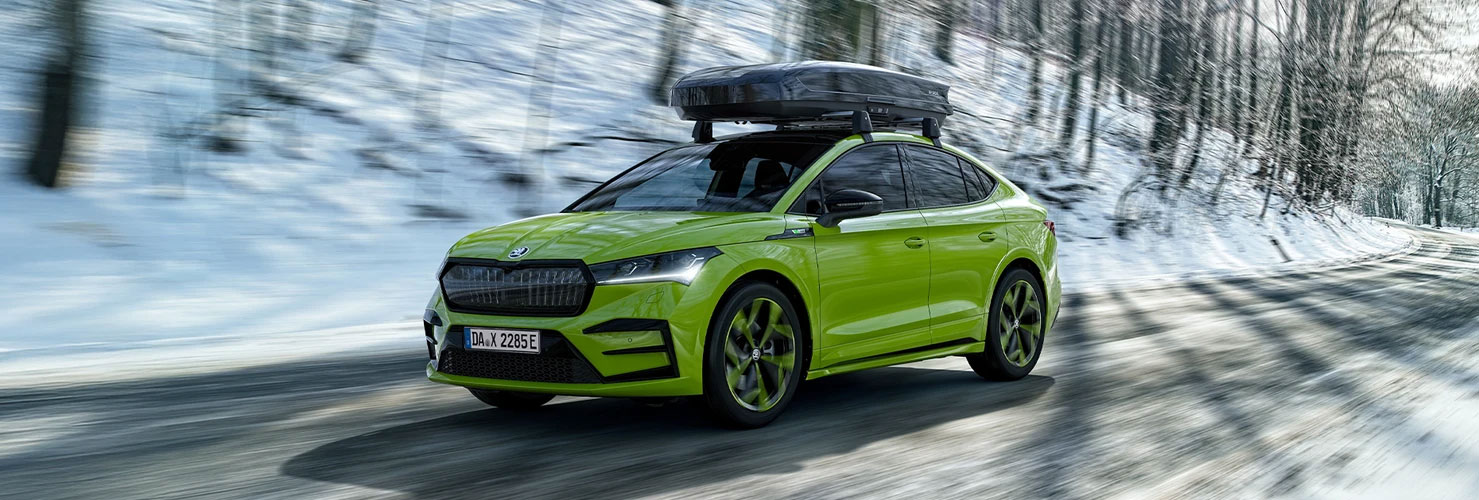 Das Skoda Enyaq in Grün mit einem Dachgepäckträger fährt schnell auf einer verschneiten kurvigen Straße, umgeben von verschneiten Bäumen. Der Hintergrund zeigt einen hellen Wintertag mit Sonnenlicht. Das Skoda Enyaq in Grün mit einem Dachgepäckträger fährt schnell auf einer verschneiten kurvigen Straße, umgeben von verschneiten Bäumen. Der Hintergrund zeigt einen hellen Wintertag mit Sonnenlicht.